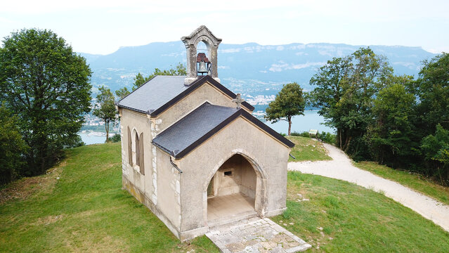 Aerial view of Notre-Dame-de-l'Etoile chapel on a grassy hill with a gravel path, surrounded by trees overlooking a lake in La Chapelle-du-Mont-du-Chat, Auvergne-Rhone-Alpes, France.