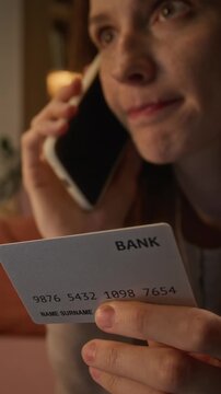 Vertical video of young Caucasian woman holding bank credit card while speaking on smartphone. Footage suits banking security apps, fraud protection, financial customer service campaigns