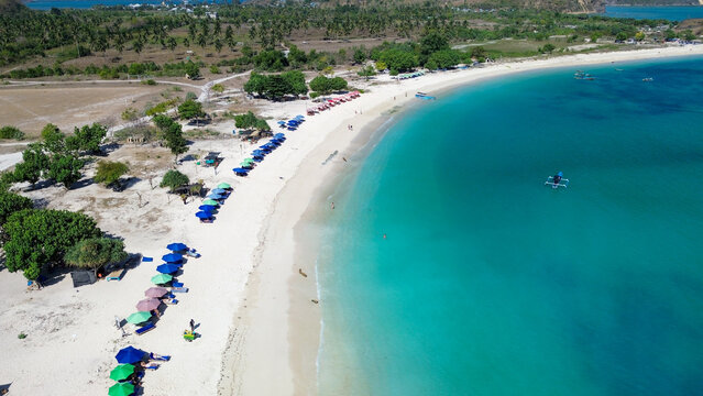 Aerial view of Tanjung Aan Beach with its turquoise waters, white sand, rows of colorful umbrellas, and lush palm trees in Central Lombok Regency, West Nusa Tenggara, Indonesia.