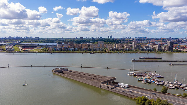 Aerial view of the IJ-tunnel entrance emerging from the water with a marina, cargo ship, and the Amsterdam skyline under a blue sky in Amsterdam, Noord-Holland, Netherlands.