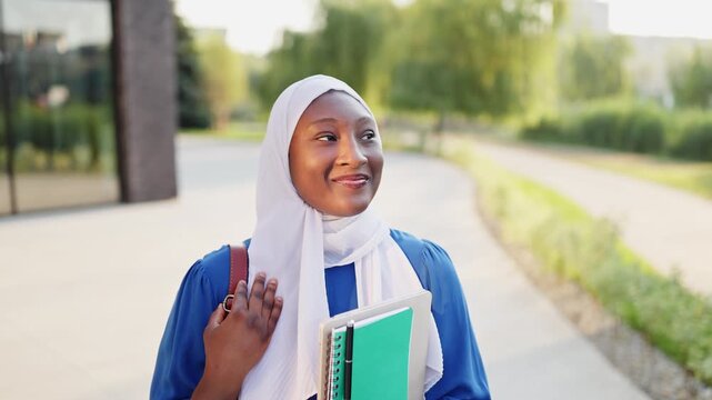 A smiling African American Muslim woman wearing a hijab and blue dress holds books while walking outdoors near a modern building
