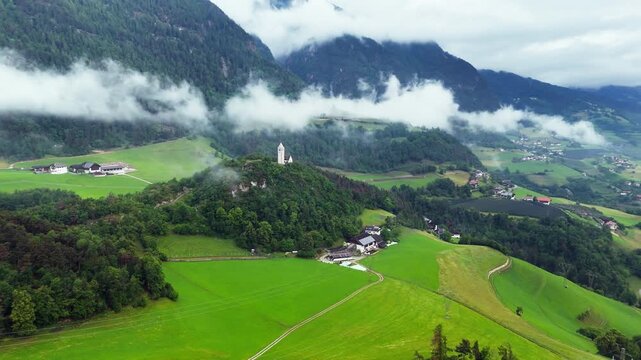Church of Saint Verena above meadows and tree lines under cloud bands, aerial view. Dolomites, South Tyrol, Italy