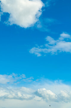 Blue sky background with scattered white cumulus clouds. A small bird flying at the bottom of the vertical frame in the distance