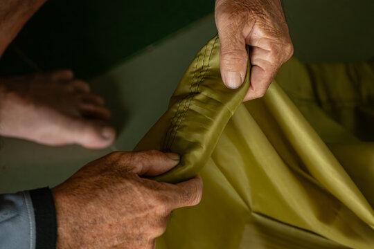  Close-up of elderly craftsman hands handling green nylon fabric hem during manual sewing process