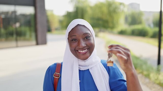 A smiling African American Muslim woman wearing a white hijab and blue dress holds a set of keys with a house charm