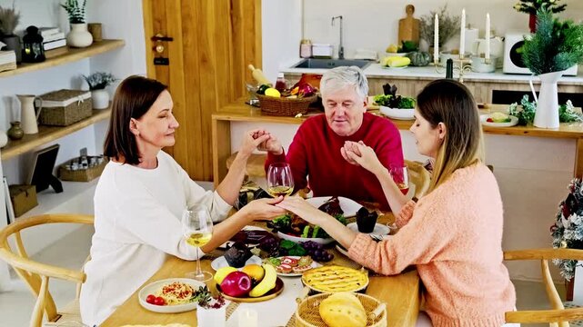 Three people hold hands in prayer before a festive meal in a cozy kitchen, surrounded by pies, fruit, and wine, sharing a warm family moment
