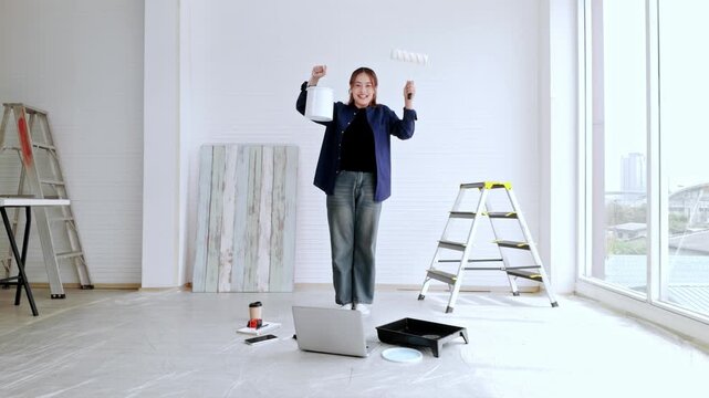 Person celebrates a home painting project in a bright room with ladders, a roller, paint tray, laptop, and big windows, suggesting a successful DIY renovation.