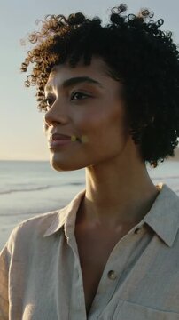 Vertical video: Turning in breeze woman in beige shirt tucking hair at shoreline, ocean lens flare
