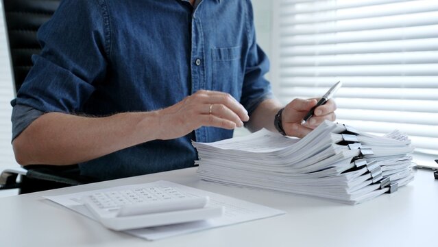 Individual working diligently on financial documents, calculating expenses and reviewing reports at an office desk, managing a large stack of paperwork and invoices