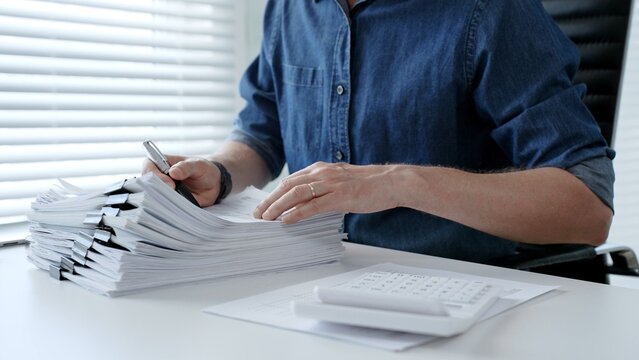 Individual working diligently on financial documents, calculating expenses and reviewing reports at an office desk, managing a large stack of paperwork and invoices