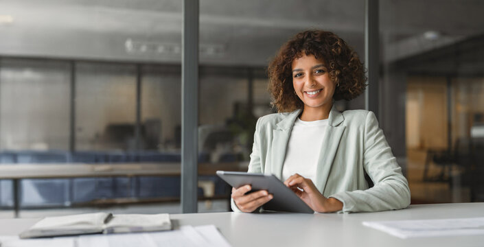 Smiling latina business woman coordinator sitting at desk, using digital tablet beside paper documents. Organized professional reviews project details with ease. Banner, copy space.