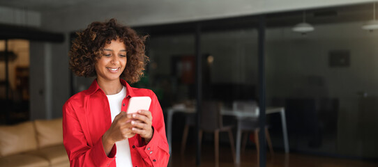 Young Latin woman using smartphone phone, texting with smile in modern office lounge. Casual...