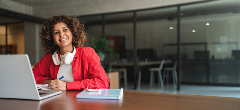 Young woman freelancer writing in notebook with pen beside open laptop, casual style at office desk. Curly haired creative jots down ideas during relaxed work session. Banner, copy space.