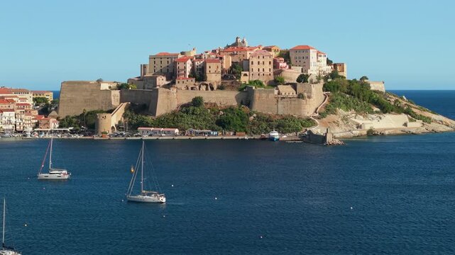 Aerial 70mm telephoto drone view of Calvi Citadel with sailboats in Corsica France. Luxury sailing yachts anchored near historic peninsula 4K