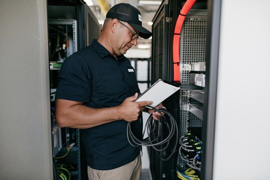 Technician in a data center holding a blank tablet mockup and network cables, inspecting grainy server racks for network maintenance and IT support