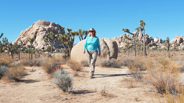 Mature woman wearing hat walking on dirt trail, enjoying scenic desert landscape of joshua tree national park during a sunny day trip, exploring the unique rock formations