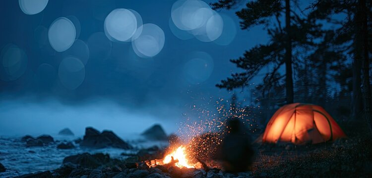 Orange tent glows at night beside a campfire on a misty, rocky shore