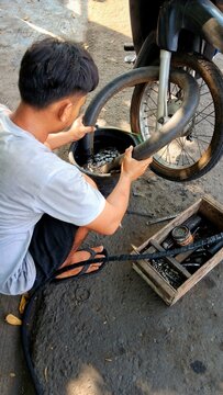A mechanic crouches beside a motorcycle, holding an inner tube over a bucket of liquid, with tools, spare parts, and the front wheel visible on concret