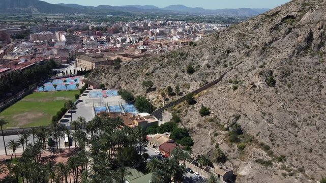 Smooth drone shot flying past rugged mountains toward the historic city of Orihuela in Alicante, Spain. Scenic aerial cityscape view under bright sunlight.