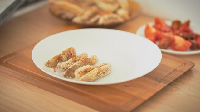 Oven-baked eggplant transferred from a baking tray to a white plate by a housewife using tongs in daylight. Concept of healthy homemade cooking and natural oven-baked food preparation