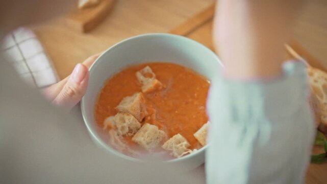 Overhead shot of a woman enjoying homemade tomato soup with croutons and a wooden spoon in a daylight home kitchen. Theme of healthy natural food and warm domestic cooking at home