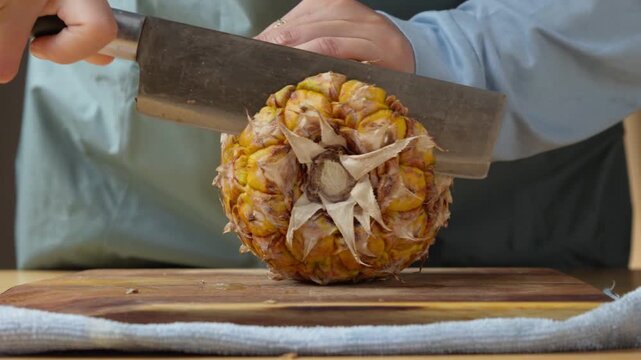 Person cutting off the top crown of a fresh ripe pineapple using a large kitchen knife on a wooden cutting board