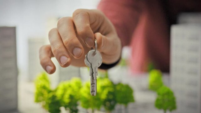 A close-up of shiny keys held out by a man's hand against a backdrop of miniature modern residential complexes. Conceptualization of real estate investment and purchasing a home in a new building