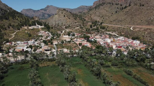 Aerial descending drone shot of San Ant&oacute;n village nestled in the lush Orihuela palm grove, surrounded by arid desert mountains in Alicante, Spain.