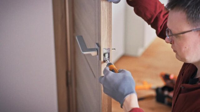 A close-up of gloved hands tightening a door handle mechanism with an orange screwdriver on a wooden interior door with an orange toolbox in the background. Concept of professional lock repair