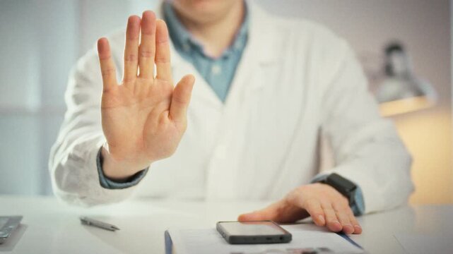 A hand raised by a doctor in a white coat signals a clear stop gesture, palm outward, in a close-up medical office scene. This universal medicine cue conveys warning, control, and patient safety
