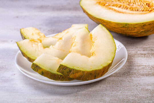 Sliced piel de sapo melon on white plate with half melon in background, green rind and pale orange flesh, rustic textured surface