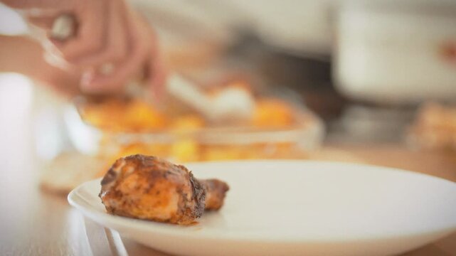A housewife plates roasted meat and potatoes using metal tongs onto a white dish in close-up. Home kitchen in daylight. Concept of Thanksgiving celebration and festive homemade cooking