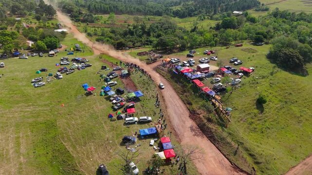 Rally campsite and spectators lining a dirt road in Nueva Alborada, Paraguay, aerial orbit as dust rises behind speeding racecar
