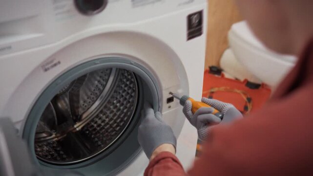 A specialist in work uniform and gloves disassembles a washing machine body in a bathroom next to a toolbox and toilet. Concept of reliable repair and maintenance of domestic home appliances