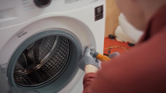 A technician in gloves carefully removes fixing bolts from the body of a washing machine with a drum window in a bathroom with a toolbox in the background. Concept of technical diagnostics and repair