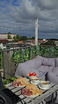 Breakfast table served with coffee, fresh bread, cold cuts, and fruit bowl on a rooftop terrace in Rostock with gray cushions and city roofs view.