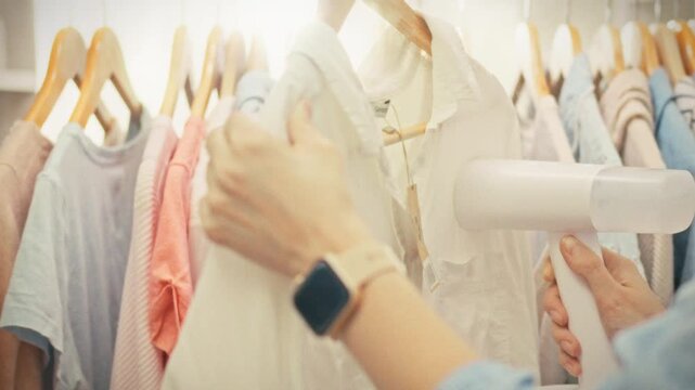 A woman uses a handheld steamer to smooth a garment in a clothing store surrounded by multiple items hanging on display racks. Concept of professional care and maintenance of clothing in a shop