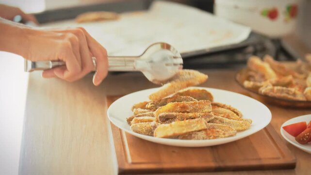 Baked eggplant. A housewife transfers oven-baked eggplant from a baking tray to a white plate using tongs. Home kitchen in daylight. Concept of healthy oven-baked homemade cooking