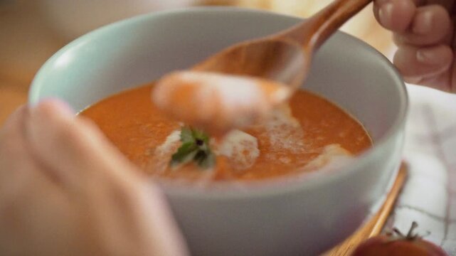 Close-up of warm homemade tomato soup with croutons in a blue bowl. A woman eats with a wooden spoon beside fresh bread on a white towel. Concept of healthy autumn harvest meal