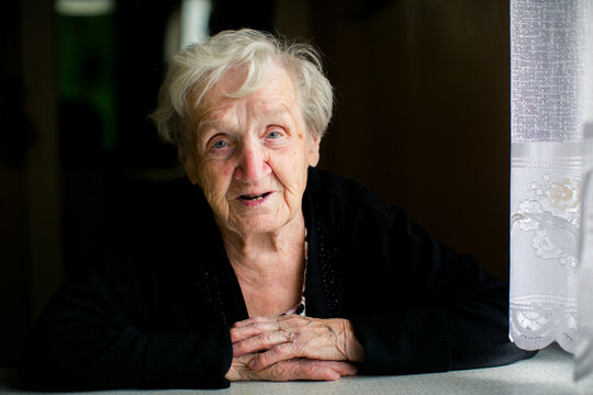 Portrait of an elderly woman resting her folded hands on a table by a window and looking at the camera indoors