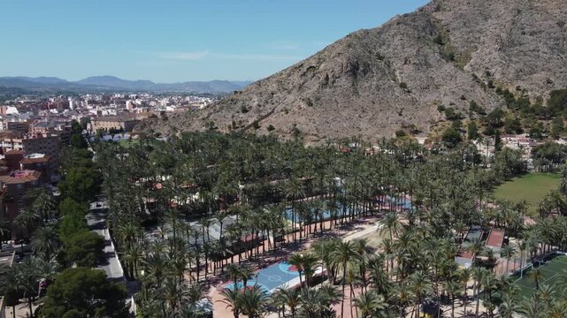 Aerial view flying over a lush palm grove and sports fields toward the historic San Anton chapel, with the majestic Orihuela mountain range rising in the background.