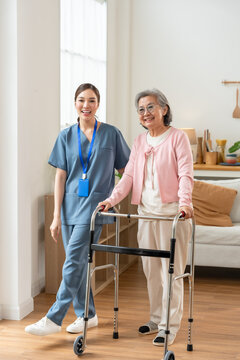 Senior Asian woman stands with walker beside young Asian nurse during rehabilitation, supporting mobility, balance, confidence and independence inside modern assisted living senior care center.