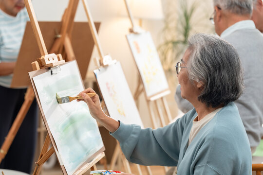 Senior Asian woman painting on canvas during creative art activity with other elderly residents in assisted living community, supporting mindfulness, social engagement, healthy aging and wellbeing.