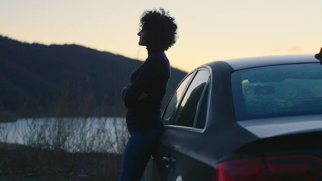A woman stands leaning next to a black luxury car looking at sunset over mountain lake. The sky is orange and the sun is setting