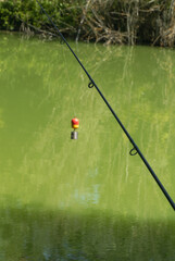Fishing rod with float and feeder hanging above calm green pond water in quiet freshwater angling scene © Miroslav