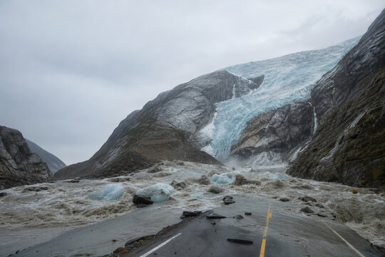 Alpine glacier collapse flooding mountain road with submerged cracked highway and rushing ice debris illustrating climate change infrastructure vulnerability and glacial flood risk emergency