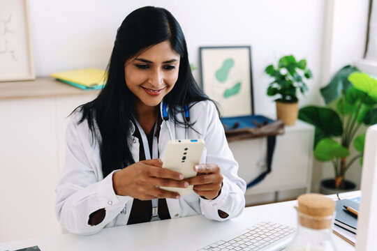Smiling Indian female doctor looking at smartphone in bright clinic office. Friendly healthcare professional reading message on phone at desk. Healthcare and technology concept.