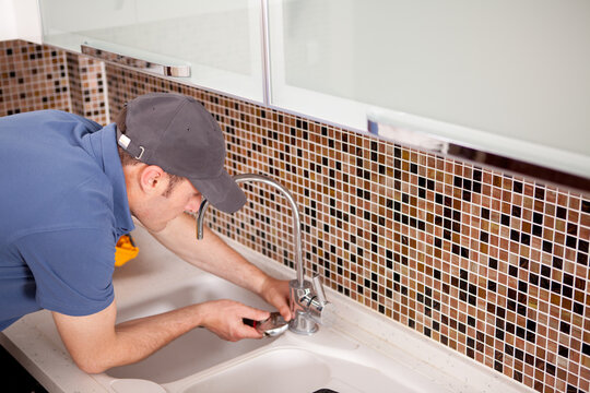 Plumber wearing cap fixing kitchen faucet with tools in modern kitchen while bending over sink.