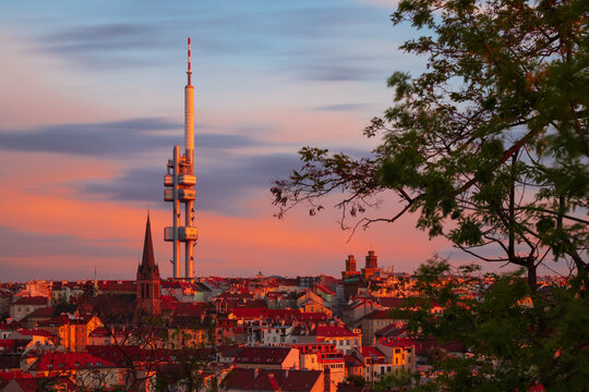 Žižkov Television Tower rising above Prague at sunset, with St. Procopius Church and red-roofed cityscape glowing in warm evening light, Czech Republic.