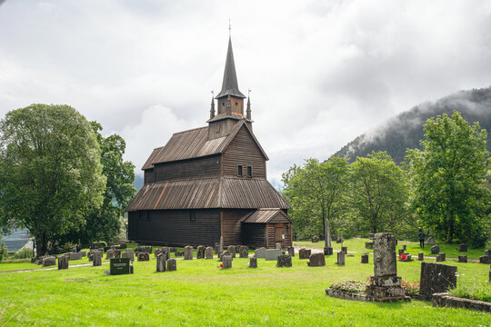 Historic dark wooden stave church stands in a green cemetery with ancient headstones under a cloudy sky with misty mountains in the background in Norway.